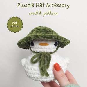 Pode incluir: Um boneco de neve de pelúcia de crochê usando um chapéu e cachecol verdes. O boneco de neve é branco com olhos pretos e um bico laranja. A imagem inclui o texto "Plushie Hat Accessory crochet pattern" e "PDF pattern".