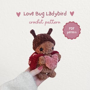 Könnte beinhalten: Gehäkeltes Marienkäfer-Spielzeug mit braunem Kopf und Körper, das ein rosa Herz hält. Es hat einen roten Umhang und Antennen. Der Text "Love Bug Ladybird crochet pattern" steht über dem Spielzeug. PDF-Muster verfügbar.