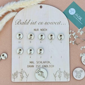 Peut inclure: Un tableau de compte à rebours en bois avec le texte "Bald ist es soweit..." et "NUR NOCH". Le tableau comporte 10 petits cercles avec des motifs floraux, chacun avec un numéro de 1 à 10. Le tableau comporte également le texte "MAL SCHLAFEN, DANN IST ENDLICH" et un petit cercle avec un motif floral et un petit sac avec une étoile.