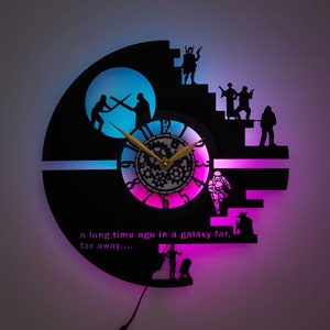 May include: A black and silver wall clock shaped like the Death Star from Star Wars. The clock has a pink and blue light behind it and the text "A long time ago in a galaxy far, far away..." is printed on the clock.