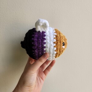 May include: A crocheted bee plush toy with purple, white, and brown stripes. The bee has a small white crocheted ear.
