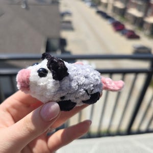 May include: A handmade crochet opossum toy. The plush animal features a white face with pink nose and ears, black eyes and ears, and a gray and white body with black spots. The tail is pink.