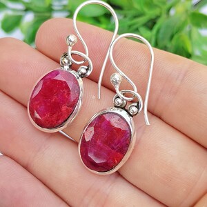 May include: A pair of silver earrings with oval, deep red gemstones. The gemstones are set in a silver bezel and suspended from a decorative silver hook. The earrings are displayed against a neutral background.