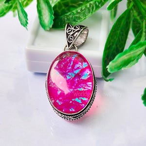 Puede incluir: Un colgante de plata con una piedra de ópalo rosa. El ópalo tiene un brillo iridiscente con matices azules y morados.