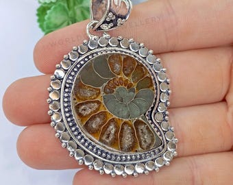 May include: A silver pendant featuring a fossilized ammonite with a spiral pattern in shades of brown, green, and white. The pendant is surrounded by a decorative border of silver beads.