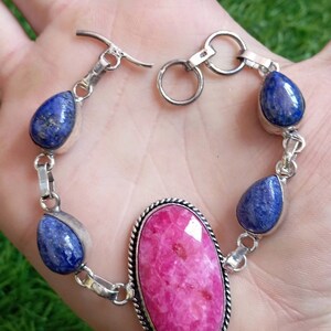 May include: A silver bracelet featuring a large oval pink stone and smaller teardrop-shaped blue stones. The bracelet has a silver clasp.