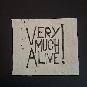 May include: A square, off-white fabric patch with the words "VERY MUCH ALIVE!" printed in black, block letters. The patch has a raw, unfinished edge and is set against a black background.