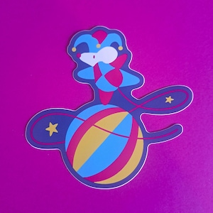 Puede incluir: Una pegatina troquelada que muestra un ratón blanco con un sombrero de bufón y un traje a juego en azul y rosa. El ratón está equilibrado sobre una gran pelota redonda multicolor con rayas amarillas, azules y rosas, rodeada de dos estrellas amarillas.