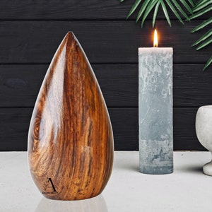 Puede incluir: Una urna de madera en forma de l&aacute;grima con un acabado de grano de madera marr&oacute;n oscuro. La urna est&aacute; sentada sobre una superficie blanca con una vela gris y un mortero y un pil&oacute;n blancos en el fondo.