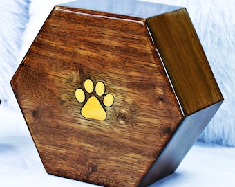 May include: A hexagonal wooden pet urn with a gold paw print design. The urn is made of dark wood with a glossy finish. The text "Art Galore Valley" is inscribed on the bottom of the urn.