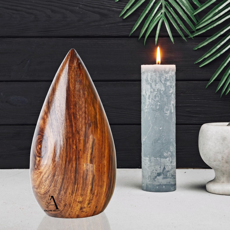 Puede incluir: Una urna de madera en forma de l&aacute;grima con un acabado de grano de madera marr&oacute;n oscuro. La urna est&aacute; sentada sobre una superficie blanca con una vela gris y un mortero y un pil&oacute;n blancos en el fondo.