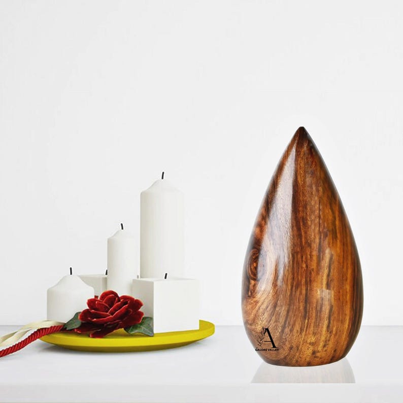 Puede incluir: Una urna de madera con forma de l&aacute;grima, con un acabado de madera de grano marr&oacute;n oscuro. La urna est&aacute; sentada sobre una superficie blanca, frente a un fondo blanco. Hay cuatro velas blancas y una rosa roja en un plato amarillo en primer plano.