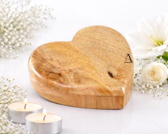 Puede incluir: Una caja de madera en forma de corazón con tapa, hecha de madera de color claro. La caja está rodeada de flores blancas y dos velas de té encendidas. La caja tiene un acabado de veta de madera natural.