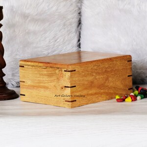 Puede incluir: Una caja de madera con un acabado de madera natural y molduras de color marrón oscuro. La caja está cerrada y se encuentra sobre una superficie blanca. El texto "Art Galore Valley" está impreso en la parte delantera de la caja.
