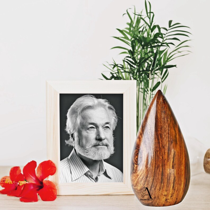 Puede incluir: Una urna de madera con forma de l&aacute;grima, con veta de madera marr&oacute;n oscuro. La urna est&aacute; sobre una mesa junto a una foto en blanco y negro enmarcada de un hombre con barba. Una flor de hibisco roja est&aacute; en primer plano.