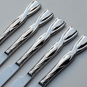 May include: Six stainless steel steak knives with a decorative twisted handle design. The blades are serrated and the handles have a polished, reflective finish. The knives are arranged diagonally on a light-colored surface.