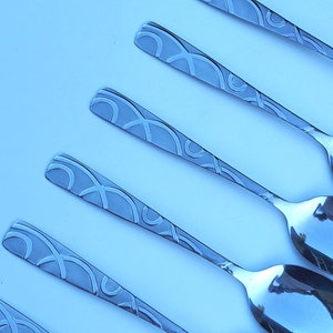 May include: Six silver spoons with a decorative pattern on the front. The spoons are arranged in a row, with the handles pointing to the right.