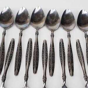May include: A set of ten silver spoons with ornate designs on the handles. The spoons are arranged in two rows, with five spoons in each row.