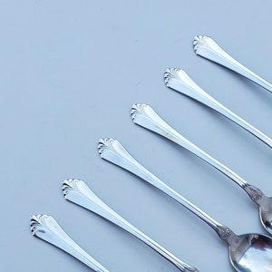 May include: Six silver spoons arranged diagonally. Each spoon has a long, slender handle and a rounded bowl. The handles have a decorative floral design near the bowl. The spoons are reflective, showing the surrounding environment.