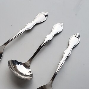 May include: Three silver spoons with ornate handles. The spoons are on a white background. The spoon on the left has a slotted bowl, the middle spoon has a rounded bowl, and the spoon on the right has a rounded bowl.