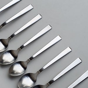 May include: A set of eight silver teaspoons arranged in a row on a white surface. The spoons are all the same size and shape, and they have a simple, elegant design.