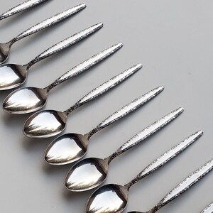 May include: A set of eight silver spoons with an ornate design. The spoons are arranged in a fan shape, with the handles pointing towards the top of the image.