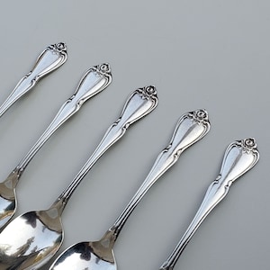 May include: Five silver teaspoons with ornate handles. The spoons are arranged in a row, with the handles pointing to the right.