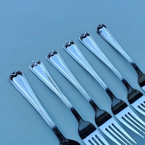 May include: Six silver forks with ornate handles, arranged in a row on a light blue background.