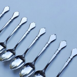 May include: A set of eight silver teaspoons with ornate handles. The spoons are arranged in a row, with the handles pointing towards the right side of the image.
