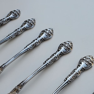 May include: Six silver ornate spoons with decorative handles. The spoons are arranged in a row, with the handles pointing to the right.