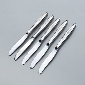 May include: Five silver butter knives with ornate designs on the handles.