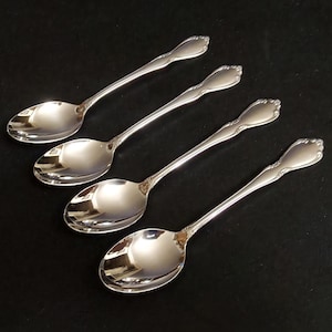 May include: Four silver spoons with a reflective surface are arranged diagonally on a black background. Each spoon has a long, slender handle and a rounded bowl. The handles have a decorative, scalloped design at the end.