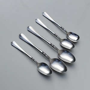 May include: Five silver teaspoons with ornate handles. The spoons are arranged in a row, with the handles pointing to the right. The spoons are all the same size and shape.