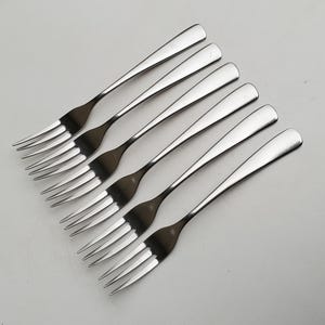 May include: Six stainless steel forks arranged diagonally on a white surface. Each fork has a long, slender handle and four tines. The forks are reflective, with a polished finish, and are evenly spaced.