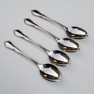 May include: Four silver spoons with ornate handles. The spoons are arranged in a row, with the handles pointing to the right. The spoons are polished and shiny.