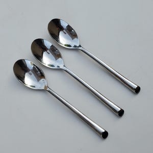 May include: Three shiny, stainless steel spoons with oval bowls and long, slender handles. The spoons are arranged diagonally, reflecting light and creating a bright, clean aesthetic. The spoons are a kitchen utensil.