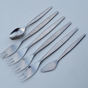 May include: A set of six silver-toned flatware pieces, including a spoon, a butter knife, and four forks. The pieces are arranged in a row on a white surface.