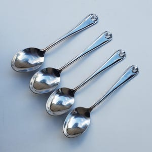 May include: Four silver spoons with a reflective surface are arranged diagonally. Each spoon has a rounded bowl and a handle with a decorative, stylized floral design at the end. The spoons are set against a light background.