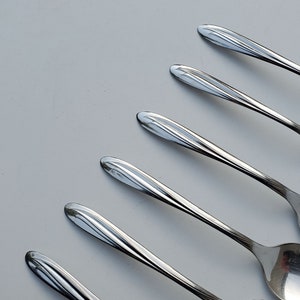 May include: Several shiny, silver spoons are arranged diagonally on a light grey surface. Each spoon features a teardrop-shaped bowl and a handle with a decorative, ridged pattern. The spoons reflect light, creating highlights and shadows.