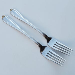 May include: Two silver forks with a decorative design on the handles. The forks are angled towards the upper right, with the tines facing down. The handles have a slight curve and a decorative element at the top.