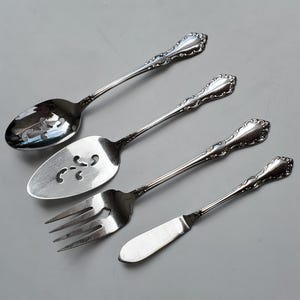 May include: Four pieces of silver-colored flatware are arranged diagonally on a gray surface. The set includes a serving spoon, a pie server, a fork, and a butter knife. Each piece has a decorative handle.