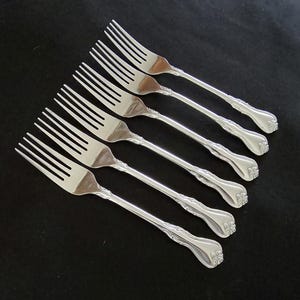 May include: Six silver forks with ornate handles are arranged diagonally on a black surface. The forks have tines and decorative detailing on the handles. The silverware is likely for formal dining.