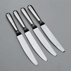 May include: Four silver butter knives with rounded handles and a black accent at the base of the blade. The knives are arranged diagonally on a gray surface, showcasing their polished finish.