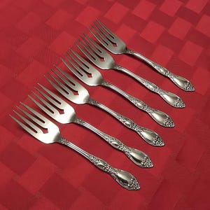 May include: Six silver forks with ornate floral detailing on the handles are arranged diagonally on a red patterned tablecloth. The forks have tines and a decorative diamond-shaped cutout.