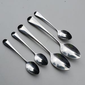 May include: Five silver spoons of varying sizes, arranged in a row on a white surface. The spoons have a rounded, oval-shaped bowl and a long, slender handle.