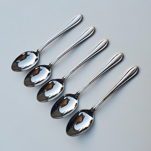 May include: Five shiny, silver spoons arranged diagonally. Each spoon has a rounded bowl and a long, ridged handle. The spoons reflect the surrounding environment, creating a mirrored effect. The spoons are made of stainless steel.