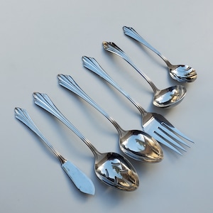 May include: A set of six silver serving utensils, including a slotted spoon, a serving spoon, a fork, and three ladles. The utensils have ornate handles and are arranged on a white surface.