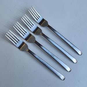 May include: A set of four identical stainless steel forks arranged in a diagonal row. Each fork features a polished metallic finish, a four-tined head, and a long, slender, flat handle that tapers slightly toward the rounded end of the utensil.