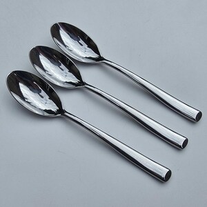 May include: Three polished stainless steel spoons arranged diagonally. Each spoon features a smooth, reflective oval bowl and a solid, slightly tapered handle with a rounded end. The metal surface shows clear reflections of the surrounding light.