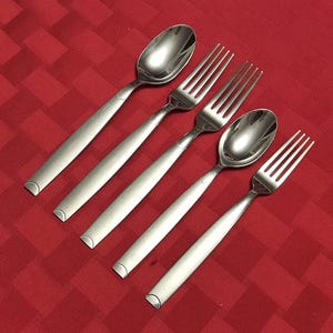 May include: A set of five stainless steel silverware pieces, including two spoons and three forks, arranged diagonally on a red tablecloth. The silverware has a simple, modern design with rounded handles and polished heads.